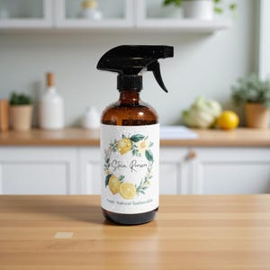 May include: An amber glass spray bottle with a black trigger nozzle. The white label features a wreath of green leaves and yellow lemons, with the text Stain Remover and Fresh Natural Sustainable printed in black script and sans-serif font.