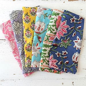 May include: A collection of six folded cloth napkins in various colors, including pink, lavender, yellow, turquoise, green, and blue. Each napkin features a different floral pattern in white, red, and green. The napkins are arranged on a white, distressed wooden surface.