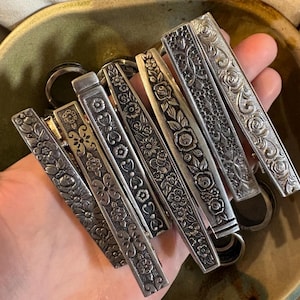 Puede incluir: Un conjunto de llaveros plateados con intrincados diseños florales y de volutas. Los llaveros rectangulares presentan varios patrones en relieve de flores, hojas y otros elementos decorativos. Están dispuestos en un cuenco de cerámica verde.