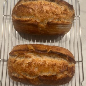 May include: Two loaves of freshly baked bread, golden brown with a crispy crust, rest on a metal cooling rack. The bread has a rustic appearance, with a textured surface and a slightly darker top.