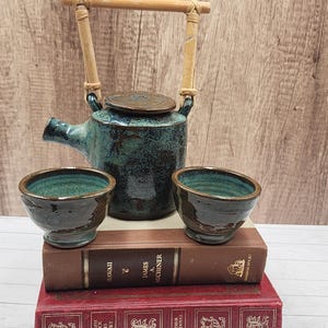 May include: A teal ceramic teapot with a bamboo handle and two matching cups. The tea set is displayed on top of two vintage books, one titled "Hawaii" and the other "The Complete Sherlock Holmes Treasury."