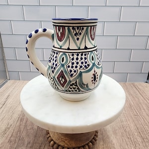 May include: A white ceramic mug with a decorative handle and a detailed pattern in blue, teal, and burgundy. The mug is adorned with geometric shapes and dots, and sits on a white marble cake stand.