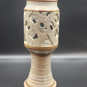 May include: A handmade ceramic candle holder with a cream-colored finish and intricate cut-out design. The holder has a cylindrical top with a decorative pattern, a pedestal base, and a brown rim. The overall height is approximately 7 inches.