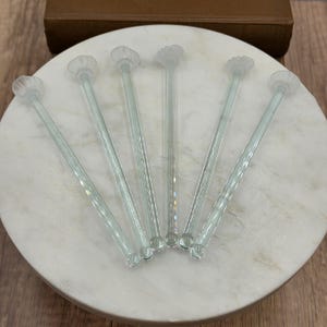 May include: A set of six clear glass cocktail stirrers, each featuring a slender, cylindrical stem and a decorative, fan-shaped top. The stirrers are arranged in a fan pattern, showcasing their transparent, smooth texture and consistent design.