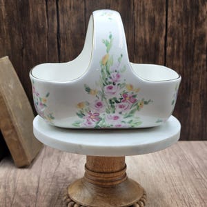 May include: A white ceramic basket with a high arched handle, featuring a hand-painted floral design of pink roses, yellow blossoms, and green leaves. The rim of the basket and the handle are accented with a thin gold trim along the edges.