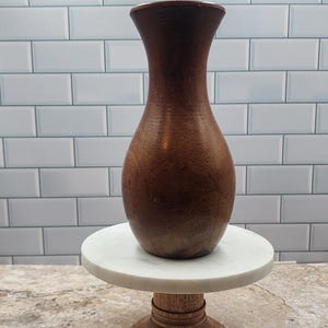 May include: A tall, brown wooden vase with a wide base and a narrow neck. The vase is displayed on a white marble-topped, wooden pedestal with a decorative beaded base. The background features a white tiled wall.