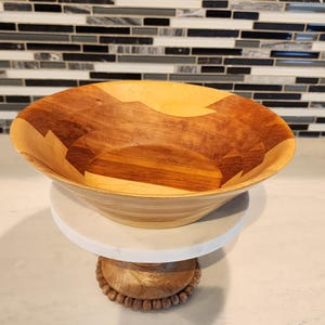 May include: A large, wooden bowl with a light and dark wood grain pattern. The bowl sits on a white marble and wood cake stand. The stand has a decorative, beaded base. The background features a mosaic tile backsplash.