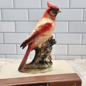 May include: A ceramic cardinal bird figurine perched on a tree branch with flowers. The bird has red, white, and brown coloring with a yellow beak. The figurine is placed on top of a brown book with the title "Hawaii".