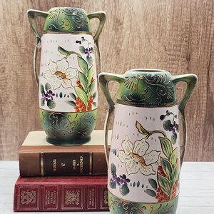 May include: Two antique ceramic vases with handles. The vases feature a light pink body with a floral design in white, green, and purple. The top and base are dark green with gold accents. The vases are displayed on top of two books.