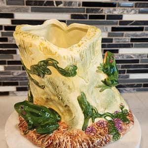 May include: A ceramic frog-themed planter. The planter is shaped like a tree stump with green frogs and vines climbing the sides. The base has brown and purple floral accents. The planter is on a white marble surface.