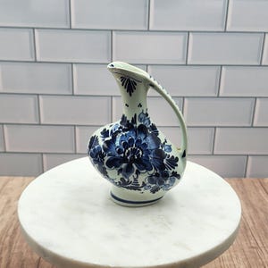 May include: A white ceramic pitcher with a bulbous body and a curved handle. The pitcher is decorated with blue floral patterns. The pitcher is sitting on a white marble surface.