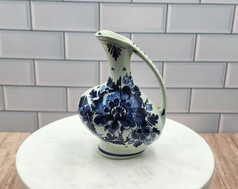 May include: A white ceramic pitcher with a bulbous body and a curved handle. The pitcher is decorated with blue floral patterns. The pitcher is sitting on a white marble surface.