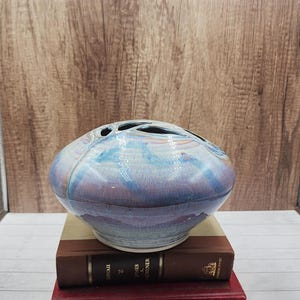 May include: A ceramic flower frog vase with a blue, purple, and white glaze. The vase has multiple holes on the top for arranging flowers. It sits atop two stacked hardcover books with gold lettering.