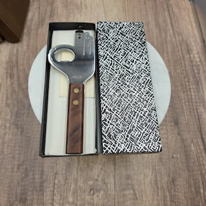 May include: A stainless steel bottle opener shaped like a hand with a wooden handle secured by two brass rivets, presented in a black box with a textured silver and black patterned lid. The metal head features a circular cutout for opening bottles.