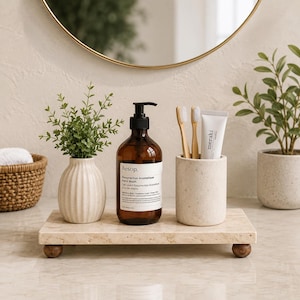 Op de afbeelding: Een rechthoekig beige stenen dienblad met vier kleine houten pootjes bevat een bruine glazen Aesop handzeepfles, een geribbelde crèmekleurige keramische vaas met een groene plant en een cilindrische stenen houder met twee tandenborstels.