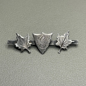 Op de afbeelding: Een zilverkleurige stropdasklem met een centraal schild met de letter "G" en twee esdoornbladaccenten. De klem is ontworpen met een staafje om aan een stropdas te bevestigen. Het metaal heeft een licht verouderd uiterlijk.