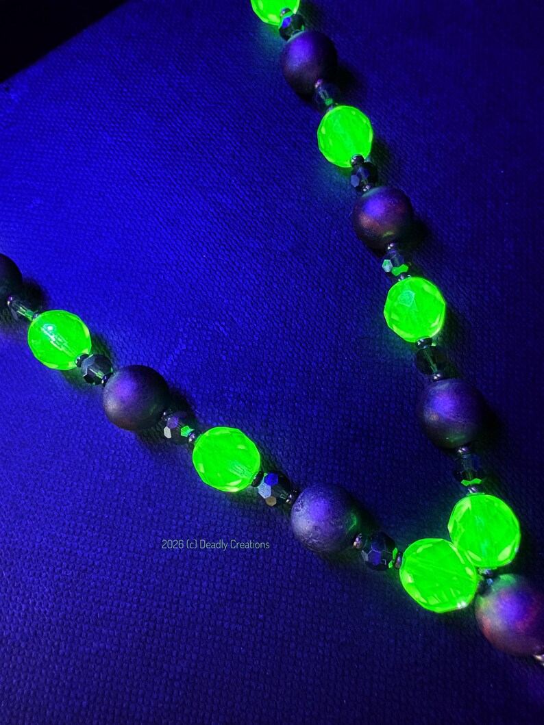 May include: A necklace featuring alternating matte dark purple spherical beads and faceted bright neon green beads that glow under ultraviolet light. The necklace includes small dark spacer beads and the text 2026 (c) Deadly Creations.