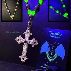 May include: A necklace featuring a silver-toned ornate cross pendant with a central dark stone, suspended from a strand of alternating matte dark beads and vibrant neon green glow-in-the-dark beads. A black display card with the text Deadly Creations.