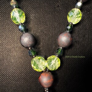 May include: A necklace featuring a repeating pattern of faceted lime green beads, dark green crystal beads, and matte brown spherical beads. The design includes a central matte brown bead flanked by two faceted lime green beads. Text reads 2026 (c) Deadly Creations.