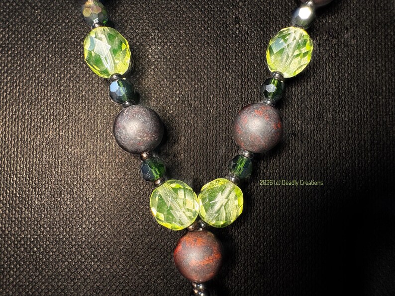 May include: A necklace featuring a repeating pattern of faceted lime green beads, dark green crystal beads, and matte brown spherical beads. The design includes a central matte brown bead flanked by two faceted lime green beads. Text reads 2026 (c) Deadly Creations.