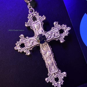 May include: A silver-toned metal cross pendant featuring intricate floral engravings and a central dark circular gemstone. The cross has ornate, filigree-style edges and is suspended from a chain. The text 2026 (c) Deadly Creations is visible.