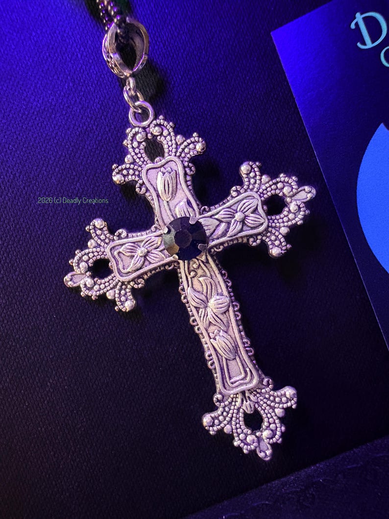 May include: A silver-toned metal cross pendant featuring intricate floral engravings and a central dark circular gemstone. The cross has ornate, filigree-style edges and is suspended from a chain. The text 2026 (c) Deadly Creations is visible.