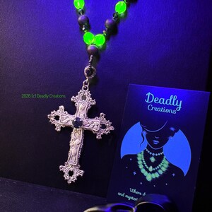 May include: A necklace featuring a silver-toned ornate cross pendant with a central dark stone, suspended from a strand of alternating matte black beads and vibrant neon green glowing beads. A black card with the text Deadly Creations is included.