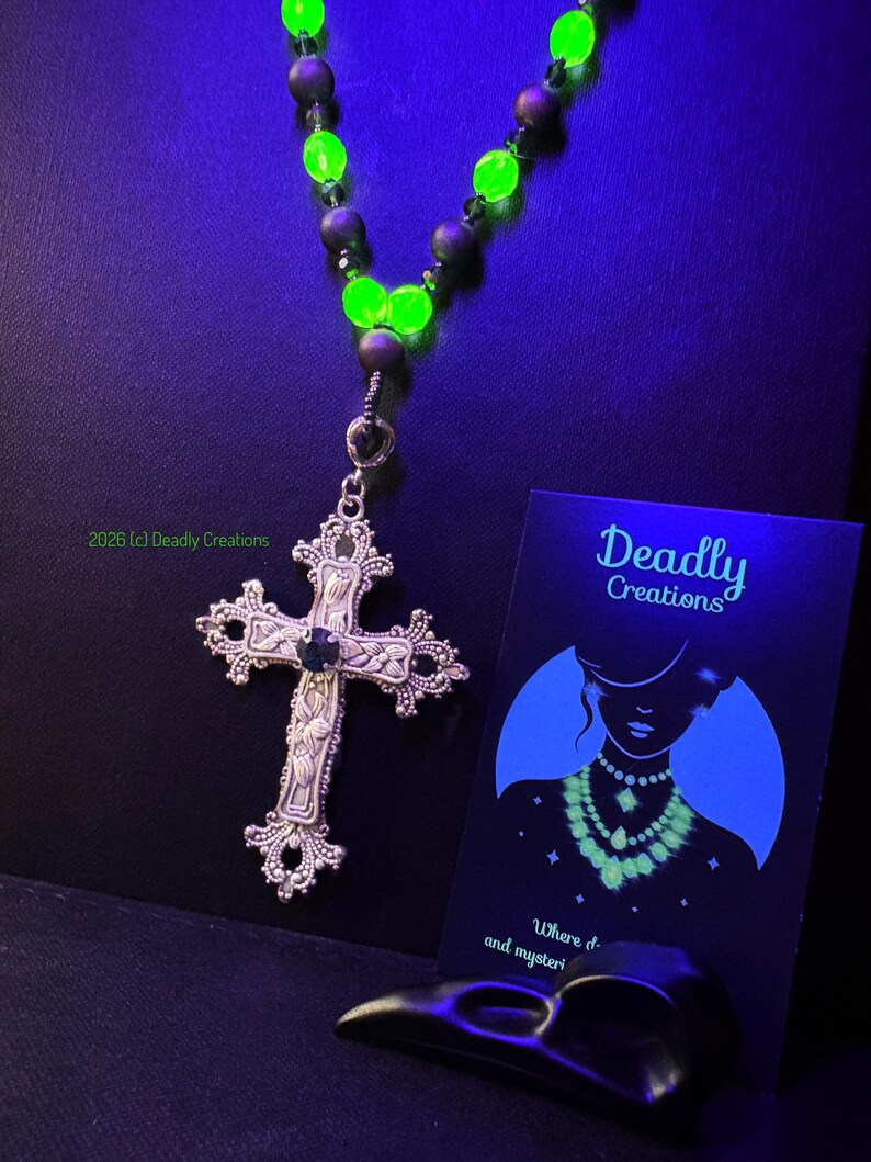 May include: A necklace featuring a silver-toned ornate cross pendant with a central dark stone, suspended from a strand of alternating matte black beads and vibrant neon green glowing beads. A black card with the text Deadly Creations is included.