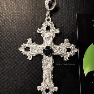 May include: A silver-toned metal cross pendant featuring intricate floral engravings and a central round black gemstone. The cross has ornate, filigree-style edges and is suspended from a dark beaded chain. The text 2026 (c) Deadly Creations is visible.