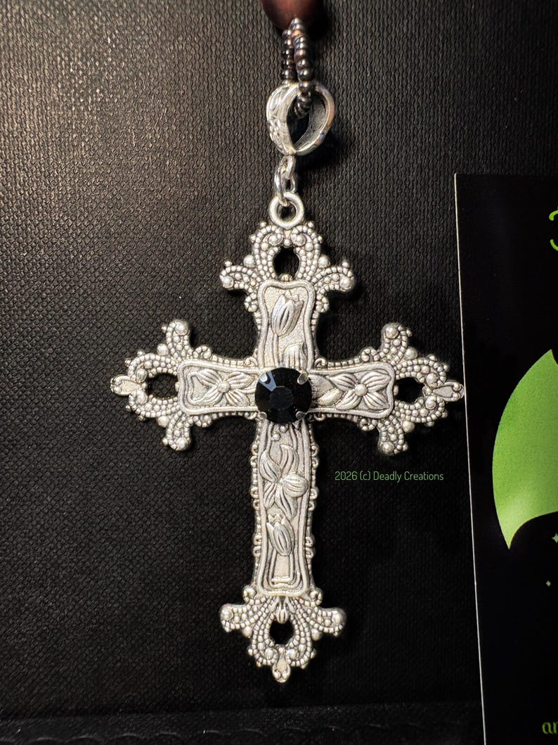May include: A silver-toned metal cross pendant featuring intricate floral engravings and a central round black gemstone. The cross has ornate, filigree-style edges and is suspended from a dark beaded chain. The text 2026 (c) Deadly Creations is visible.