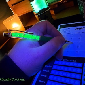 May include: A green pen with a neon green glowing interior is held over a calendar. The pen is being used to write on a calendar that says "March 2026". The image also shows a lit candle and a green glowing lantern.