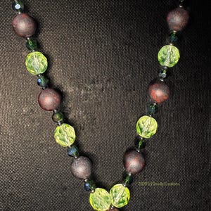 May include: A necklace featuring alternating round matte dark brown beads and faceted lime green beads, accented with small dark green spacer beads. The design includes the text 2026 (c) Deadly Creations printed in small yellow letters near the bottom.