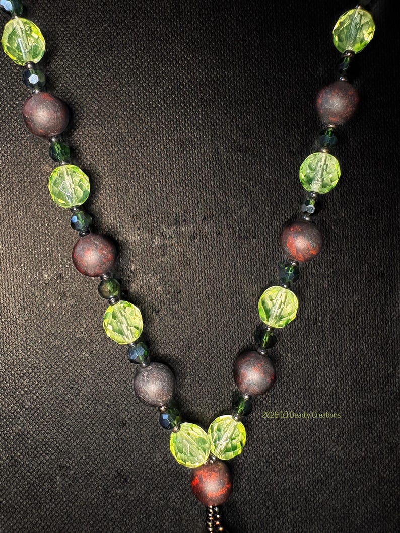 May include: A necklace featuring alternating round matte dark brown beads and faceted lime green beads, accented with small dark green spacer beads. The design includes the text 2026 (c) Deadly Creations printed in small yellow letters near the bottom.