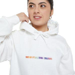 Puede incluir: Sudadera con capucha blanca con capucha con cordón y mangas largas. El texto "WEAR YOUR TRUE COLOURS" está impreso en el pecho en colores arcoíris. La sudadera con capucha está hecha de un material suave.