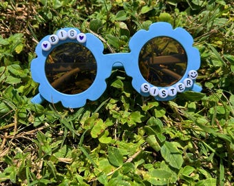 May include: Light blue flower-shaped sunglasses with brown lenses. The sunglasses have the words "BIG" and "SISTER" spelled out in white letter beads. The sunglasses are resting on a bed of green grass.