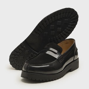 K&ouml;nnte beinhalten: Schwarze Leder-Loafer mit dicker Sohle. Der Schuh hat eine gl&auml;nzende Oberfl&auml;che und eine strukturierte, profilierte Au&szlig;ensohle. Der zweite Schuh ist umgedreht und zeigt das Sohlenmuster.