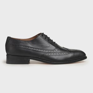 May include: A black leather Oxford shoe with brogue detailing. The shoe features a classic wingtip design with decorative perforations and a lace-up closure. The sole is a dark color, and the heel is a low block style.