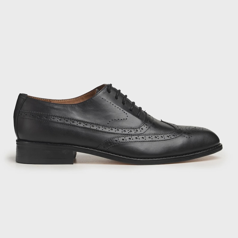 May include: A black leather Oxford shoe with brogue detailing. The shoe features a classic wingtip design with decorative perforations and a lace-up closure. The sole is a dark color, and the heel is a low block style.