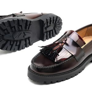 May include: A pair of burgundy leather loafers with a chunky black sole. One shoe is shown from the bottom, displaying the tread pattern. The other shoe is shown from the side, with a tassel detail.