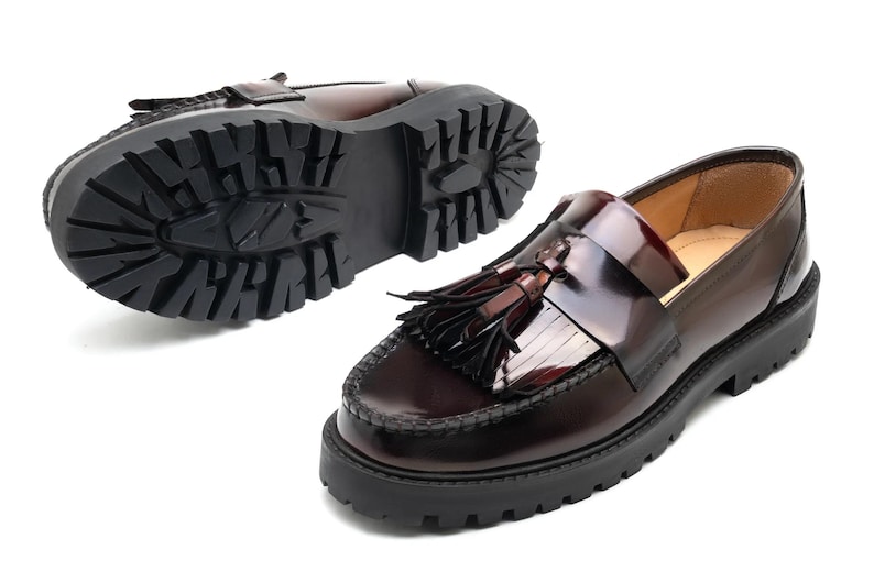 May include: A pair of burgundy leather loafers with a chunky black sole. One shoe is shown from the bottom, displaying the tread pattern. The other shoe is shown from the side, with a tassel detail.