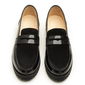 K&ouml;nnte beinhalten: Paar schwarze Leder-Loafer mit Plateausohle. Die Schuhe haben eine gl&auml;nzende Oberfl&auml;che an Zehen und Ferse, mit einem Wildleder-&auml;hnlichen Material am Blatt. Ein schwarzer Riemen verl&auml;uft &uuml;ber die Oberseite.