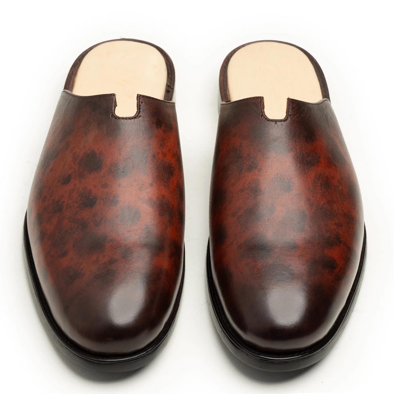 May include: A pair of brown leather slip-on shoes with a mottled pattern. The shoes have a rounded toe and a black sole. The interior is a light tan color.