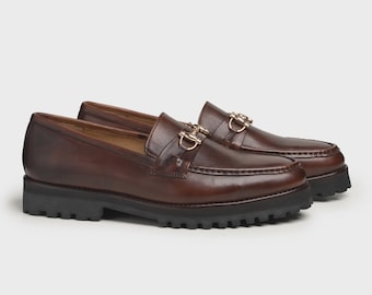 Puede incluir: Un par de mocasines de cuero marrón con detalles metálicos dorados. Los zapatos tienen un diseño clásico con punta redondeada y suela gruesa de goma negra. Los mocasines se muestran sobre un fondo neutro.