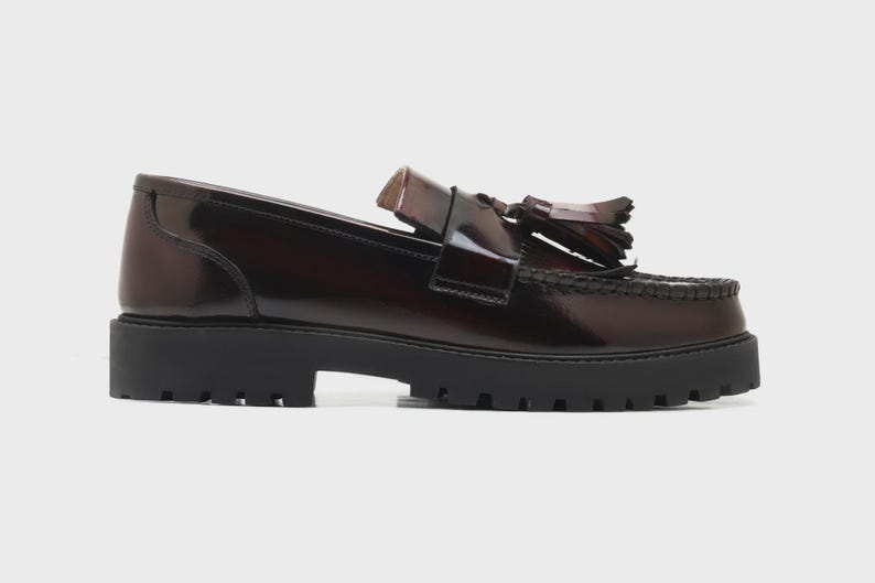 May include: A pair of brown leather loafers with a glossy finish. The shoes feature a chunky black sole and decorative tassels. The loafers have a classic design with a modern, elevated platform.