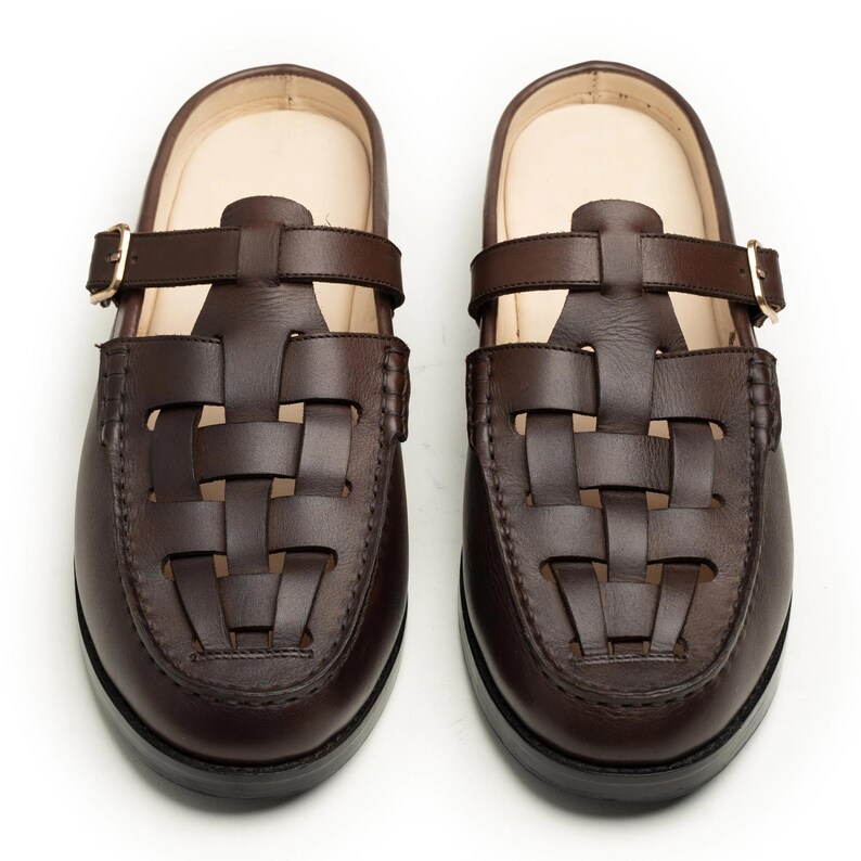 May include: A pair of brown leather clogs with a woven design on the toe and a strap with a buckle. The clogs have a dark sole and a light-colored insole.