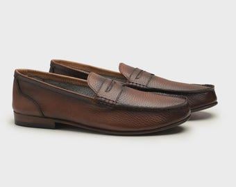May include: A pair of brown leather penny loafers. The shoes have a classic design with a rounded toe, a strap across the vamp, and a low heel. The leather has a textured appearance.