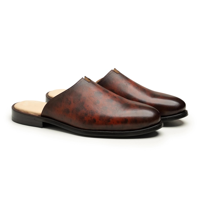May include: A pair of brown leather slip-on shoes with a dark, mottled finish. The shoes have a rounded toe, a low heel, and a black sole. The interior is a light beige color.