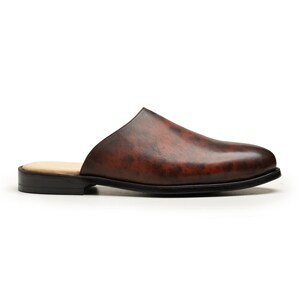 May include: A brown leather mule shoe with a rounded toe and a low, black heel. The shoe has a slip-on design and a light-colored insole. The leather has a mottled appearance, with darker and lighter areas.
