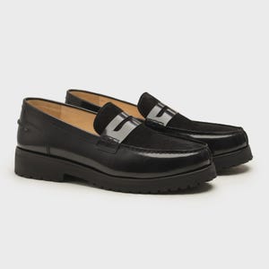K&ouml;nnte beinhalten: Ein Paar schwarze Leder-Penny-Loafer mit klobiger Sohle. Die Schuhe haben eine gl&auml;nzende Oberfl&auml;che auf dem Blatt und ein Wildleder-Panel &uuml;ber der Oberseite. Die Sohlen sind dick und die Schuhe sind vor einem neutralen Hintergrund platziert.