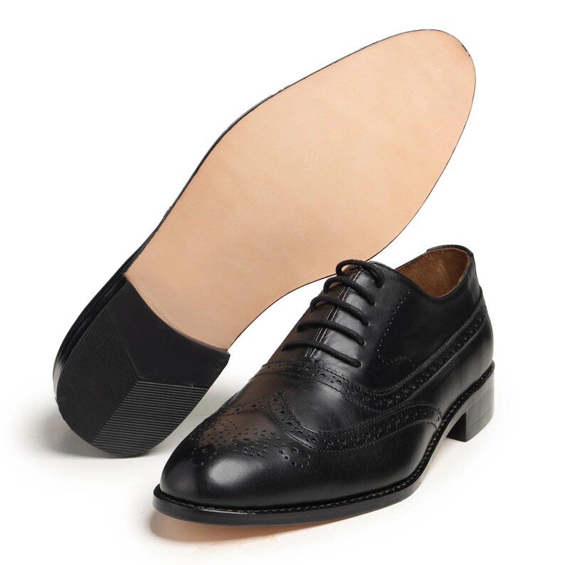 May include: Black leather Oxford dress shoe with brogue detailing and a stacked heel. The sole of a matching shoe is visible in the background, showcasing the light-colored leather sole and the dark heel.